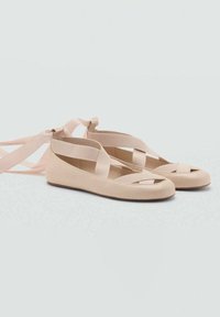 Ballerines beige pâle avec bout fermé, design découpé et brides croisées en tissu pour fermeture autour de la cheville. Texture lisse, semelle plate.