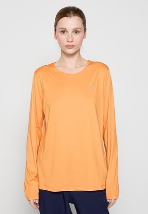 Junge Frau mit hellem Teint und zurückgebundenem Haar, die ein langärmliges orangenes Hemd und dunkle Hosen trägt, vor einem schlichten Hintergrund.