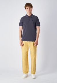 Marineblaues Poloshirt mit weißem Kragen und Akzenten, kombiniert mit gelben Hosen mit Logo-Details; das Modell steht aufrecht in weißen Sneakers.