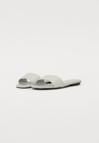 Sandales à glissière gris clair avec un bout ouvert, une sangle texturée présentant un motif, et une semelle plate. Design minimaliste et finition lisse.