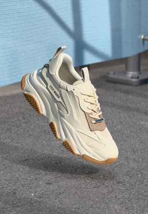 Maswny biały i beżowy sneaker z grubą, teksturowaną podeszwą i sznurowadłami, z logo "Steve Madden", zawieszony nad asfaltem obok niebieskiej ściany.