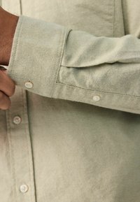 Lichtgroene shirt van zacht materiaal, met een knoopmanchet met één knoop en een gestructureerde afwerking. Zichtbare stiksels en details van de knoopsluiting.
