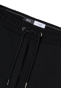 Pantalons noirs avec une taille élastique, dotés d'un cordon de serrage et d'extrémités en métal argenté. L'étiquette indique "CECI L" et "PANTS." Texture lisse.