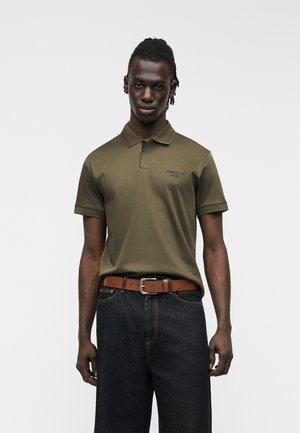 Mężczyzna w oliwkowej koszulce polo i ciemnych dżinsach z brązowym paskiem, stojący na tle jednolitego jasnoszarego tła.