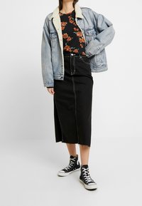Persona que lleva puesta una chaqueta de mezclilla forrada de sherpa en azul claro, una blusa negra con flores, una falda midi negra con costuras blancas y zapatillas altas negras.