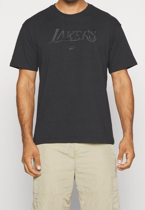 Homme portant un t-shirt noir Lakers Nike et un short cargo beige, debout les bras détendus le long du corps devant un fond uni.