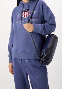Felpa blu con tasca frontale, dettagli del logo e cordini. Abbinata a pantaloni della tuta blu coordinati e una borsa di pelle nera.