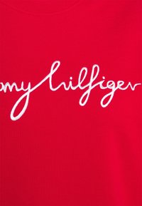 Czerwona tkanina koszulki z białym haftowanym napisem "Tommy Hilfiger" w stylu kursywy. Gładka tekstura z dekoltem typu crew neck.