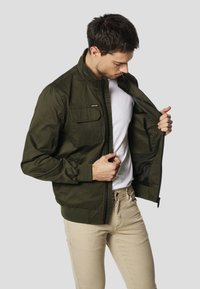 Oliwkowa kurtka bomberka z przednim zamkiem błyskawicznym i górną lewą kieszenią. Ściągacze na mankietach i dole, noszona na białej koszulce, rozpięta.