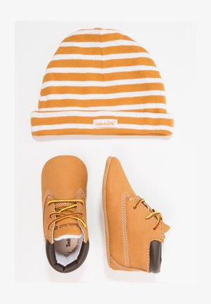 Bottines en cuir marron avec des lacets jaunes et marron, accompagnées d'un bonnet en tricot rayé orange et blanc avec une étiquette de logo.