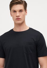 Schwarzes Baumwoll-T-Shirt mit kurzen Ärmeln, Rundhalsausschnitt und einer kleinen Brusttasche auf der linken Seite. Glatte Textur, lässige Passform, minimalistisches Design.