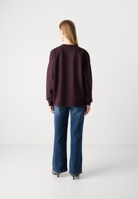 Sudadera burdeos de talla grande con cuello redondo, combinada con unos vaqueros de pierna ancha azul y calzado beige, enfatizando una silueta relajada.