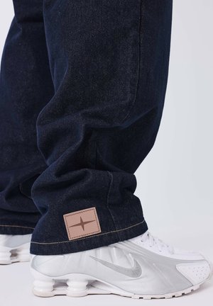 Dunkelblaue Denim-Jeans mit strukturierter Oberfläche, versehen mit einem hellbraunen Logo-Patch und einem lockeren Schnitt, kombiniert mit silbernen Nike-Sneakern.