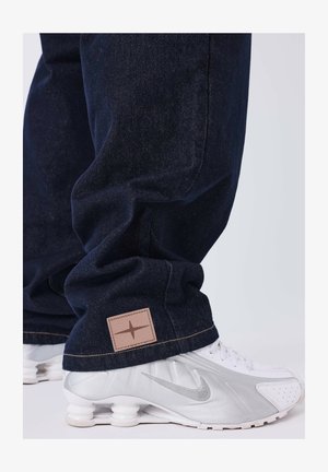 Jean en denim bleu foncé avec une finition texturée, présentant un patch logo marron clair cousu et une coupe décontractée, associé à des baskets Nike argentées.