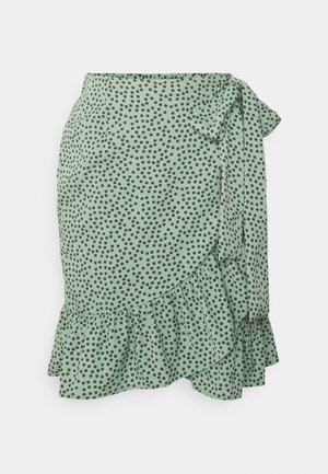 Mint green wrap skirt with black polka dots, ruffled hem, and side tie detail. Made from lightweight fabric with a soft texture.