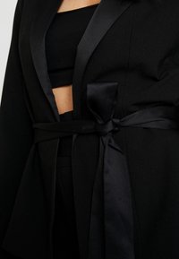 Blazer negro con solapas de satén y un lazo en la cintura. Presenta un corte entallado y un ligero escote abierto, combinado con un crop top negro debajo.