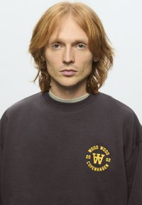 Mlada osoba s dugom crvenom kosom do ramena nosi tamni duksericu s žutim logom "Wood Wood Copenhagen 2002" na prsima.