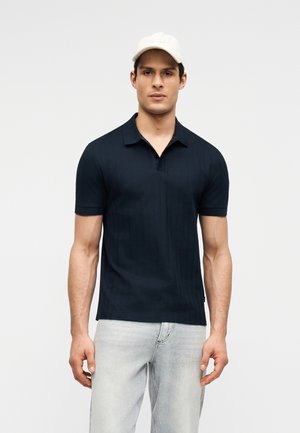 Άνδρας που φοράει μαύρο πόλο με κοντά μανίκια, ανοιχτόχρωμο τζιν και λευκό καπέλο του μπέιζμπολ, στέκεται μπροστά σε απλό φόντο.