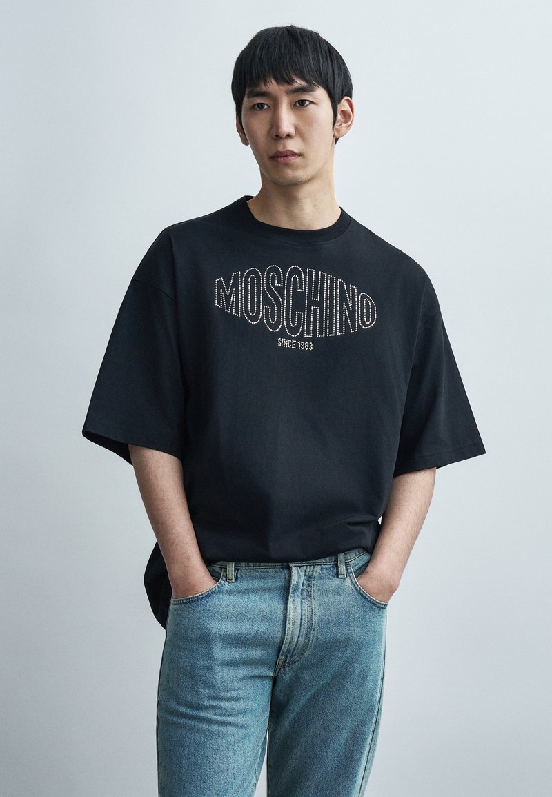 Μαύρο μπλουζάκι με κοντά μανίκια και το λογότυπο "MOSCHINO" σε λευκά γράμματα με περίγραμμα, σε συνδυασμό με ανοιχτόχρωμο τζιν παντελόνι. Διακριτική, άνετη εφαρμογή.