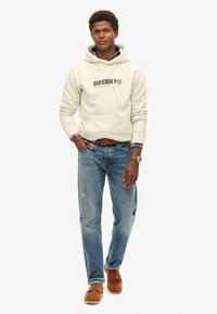 Felpa beige con logo, indossata sopra una camicia a quadretti, abbinata a jeans azzurri e scarpe marroni, con una cintura in pelle e un orologio.