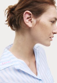 Blauw-wit gestreept shirt met een kraag; heeft roze geborduurde tekst "liefde." Meerdere gouden oorbellen in het oor. Haar in een losse knot.