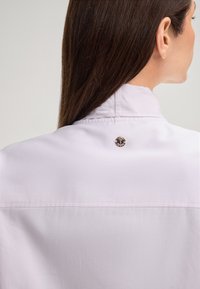 Lichtpaarse blouse met een gladde textuur en een hoge kraag, met een metalen logo-knop aan de achterkant bij de hals.