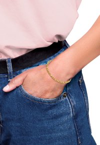 Guld kedjearmband med en länkad design, bärs på en handled, synligt mot en bakgrund av denim och en ljusrosa skjorta.