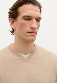 Beige V-Ausschnitt-Pullover aus einem glatten, leichten Strickmaterial. Subtile Textur mit einem kleinen bestickten Detail auf der Brust.