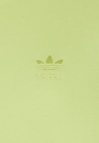 Bavlněná košile limetkově zelené barvy s malým, vyraženým logem Adidas trefoil na přední straně. Tkanina má hladký, strukturovaný povrch.