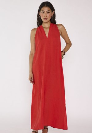 Femme debout devant un fond uni, portant une robe longue rouge sans manches avec un décolleté en V, des créoles et des sandales.