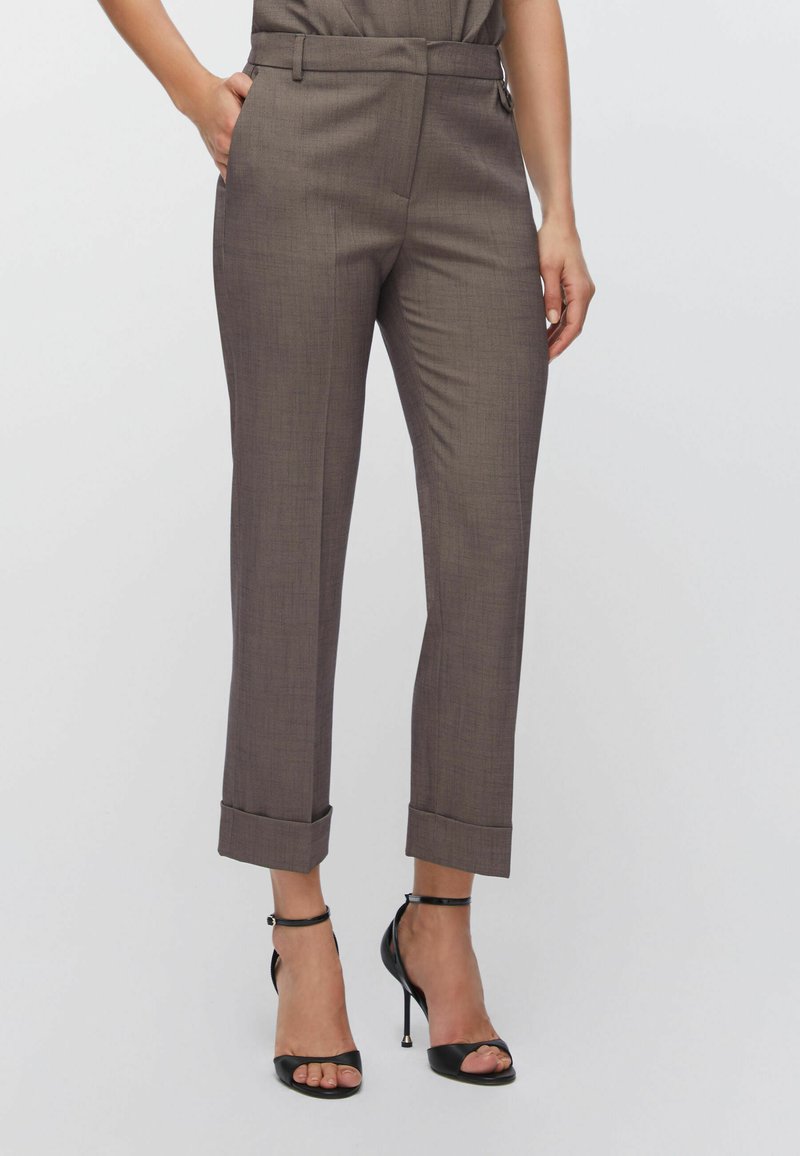 Pantalon marron sur mesure en tissu texturé, coupe ajustée, avec revers roulés. Porté avec des sandales noires à talons hauts pour créer un contraste.