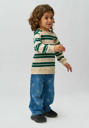 Maglione beige a righe con strisce verdi e un emblema arancione, abbinato a jeans blu con motivo a stelle e sneakers nere.