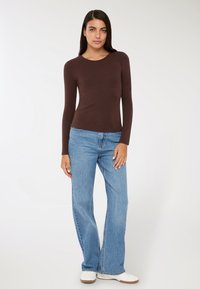 Brązowa koszula z długim rękawem zestawiona z jasnoniebieskimi spodniami dzwonami i białymi sneakersami, na neutralnym tle.