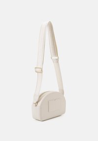 Béžová crossbody taška s kulatým tvarem, hladkým povrchem a viditelným zipem. Má jednoduchou přední kapsu a látkový popruh s logem.