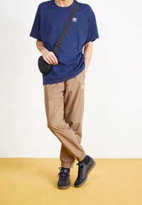 Person som bär en marinblå Adidas-t-shirt, beige Adidas-träningsbyxor, svarta sneakers och en svart axelväska, stående med benen i kors på ett gult golv.