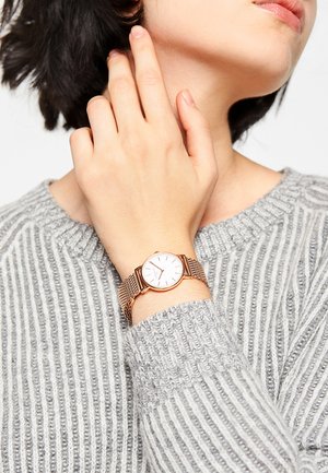 Roségoldenes Netzarmbanduhr mit weißem Zifferblatt, auf einer Hand präsentiert; getragen mit einem grauen Rippensweater. Verfügt über minimalistische Stundenmarkierungen und ein schlankes Design.