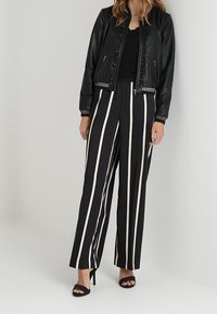 Žena v černé kožené bundě, černém topu, černých kalhotách s vysokým pasem a bílými svislými pruhy a černých sandálech na podpatku, stojí.