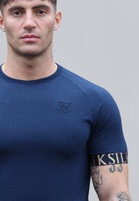 Námořnická modrá přiléhavá trička vyrobená z měkké látky s kulatým výstřihem. Obsahuje texturované logo na hrudi a kontrastní značkový manžetu.
