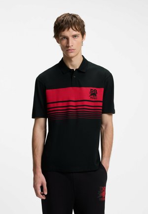 Junger Mann mit kurzem braunem Haar, der ein schwarzes Poloshirt mit roten Streifen und einem Logo auf der Brust trägt, steht vor einem einfarbigen Hintergrund.