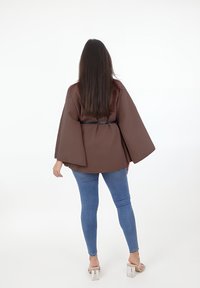 Top marrón estilo kimono con mangas anchas, ceñido a la cintura, combinado con jeans ajustados azules texturizados y sandalias de tacón bloque transparente.