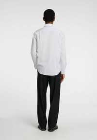 Camisa blanca de manga larga con rayas sutiles, dobladillo curvado, puños con botón, combinada con pantalones anchos negros y zapatos negros, vista desde atrás.