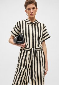 Ριγέ ολόσωμη φόρμα σε εκρού και μαύρο χρώμα με γιακά, κοντά μανίκια, κουμπιά μπροστά και ζώνη στη μέση· κρατώντας μια μικρή μαύρη τσάντα.