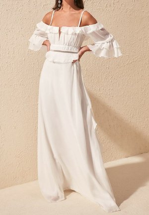 Robe blanche à épaules dénudées avec des manches à volants et une taille cintrée. Tissu léger et une longue jupe fluide avec une fente avant.