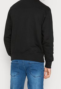Sudadera negra con cuello redondo y puños acanalados, de tela texturada, combinada con jeans de mezclilla azules con dos bolsillos traseros.