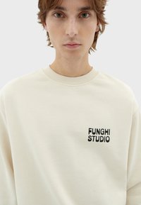 Crème kleurige sweatshirt met een ribbelboord ronde hals en subtiele zwarte geborduurde tekst "FUNGHISTUDIO" op de linkerborst. Zachte textuur.