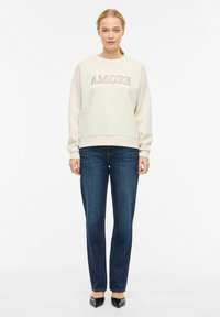 Sweat-shirt couleur crème avec le texte "AMORE" en lettres rouges, associé à un jean droit bleu foncé et des chaussures noires à bout pointu.