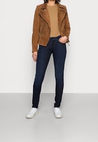 Cazadora de motociclista de gamuza marrón sobre una blusa beige, combinada con jeans ajustados oscuros y zapatillas blancas. Textura suave, diseño entallado.