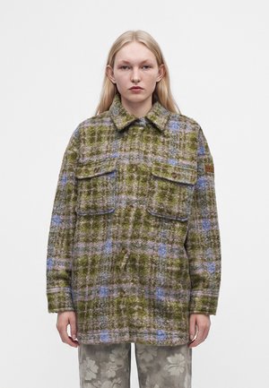 Jeune personne aux longs cheveux blonds porte une veste oversized à carreaux verts et violets avec de grandes poches et un pantalon à motif floral.