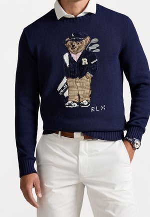 Mand iført en navyblå sweater med en golf-tema bjørn broderi, hvid skjorte med krave, hvide bukser, brun bælte og armbåndsur.