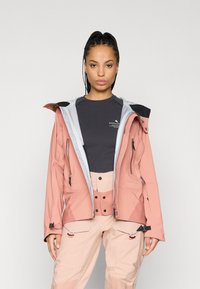 Casaco leve em tom duplo, rosa e cinza, com um design ajustado, múltiplos fechos e características ajustáveis. Usado sobre uma blusa escura e calças claras.